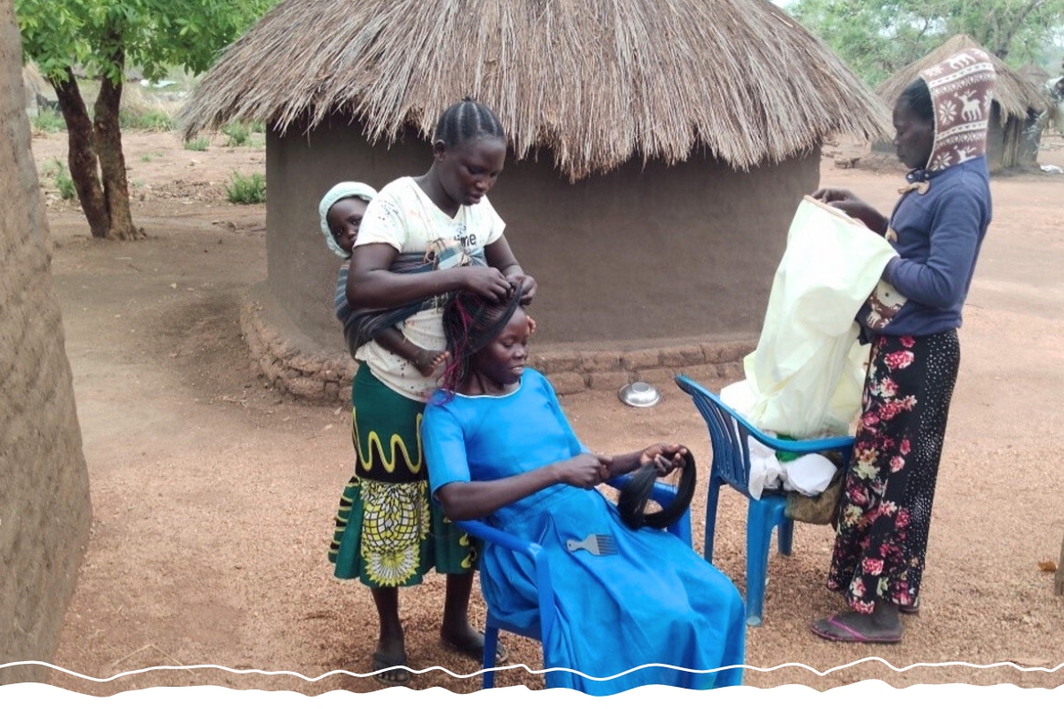 A woman with a baby on her back braids the hair of another woman sitting in a chair.